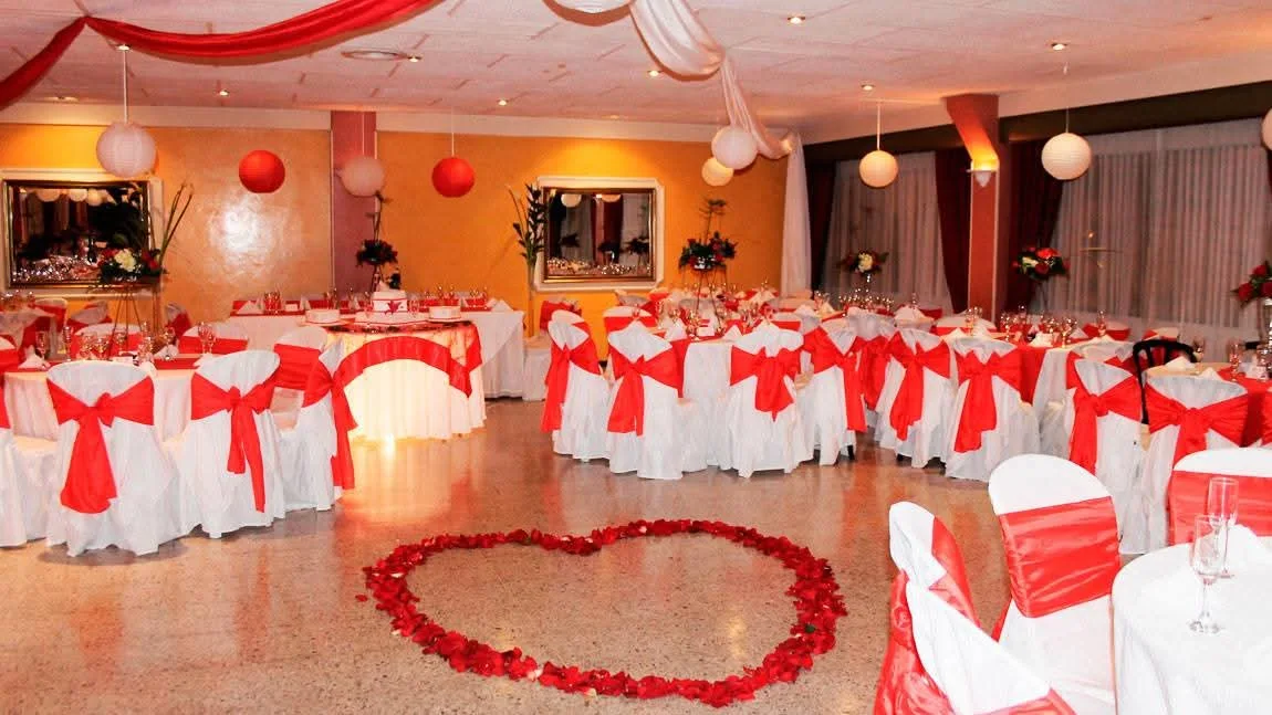 Sala de eventos decorada, con un corazón hecho de pétalos de rosas en el suelo, las mesas y sillas decoradas en blanco y rojo