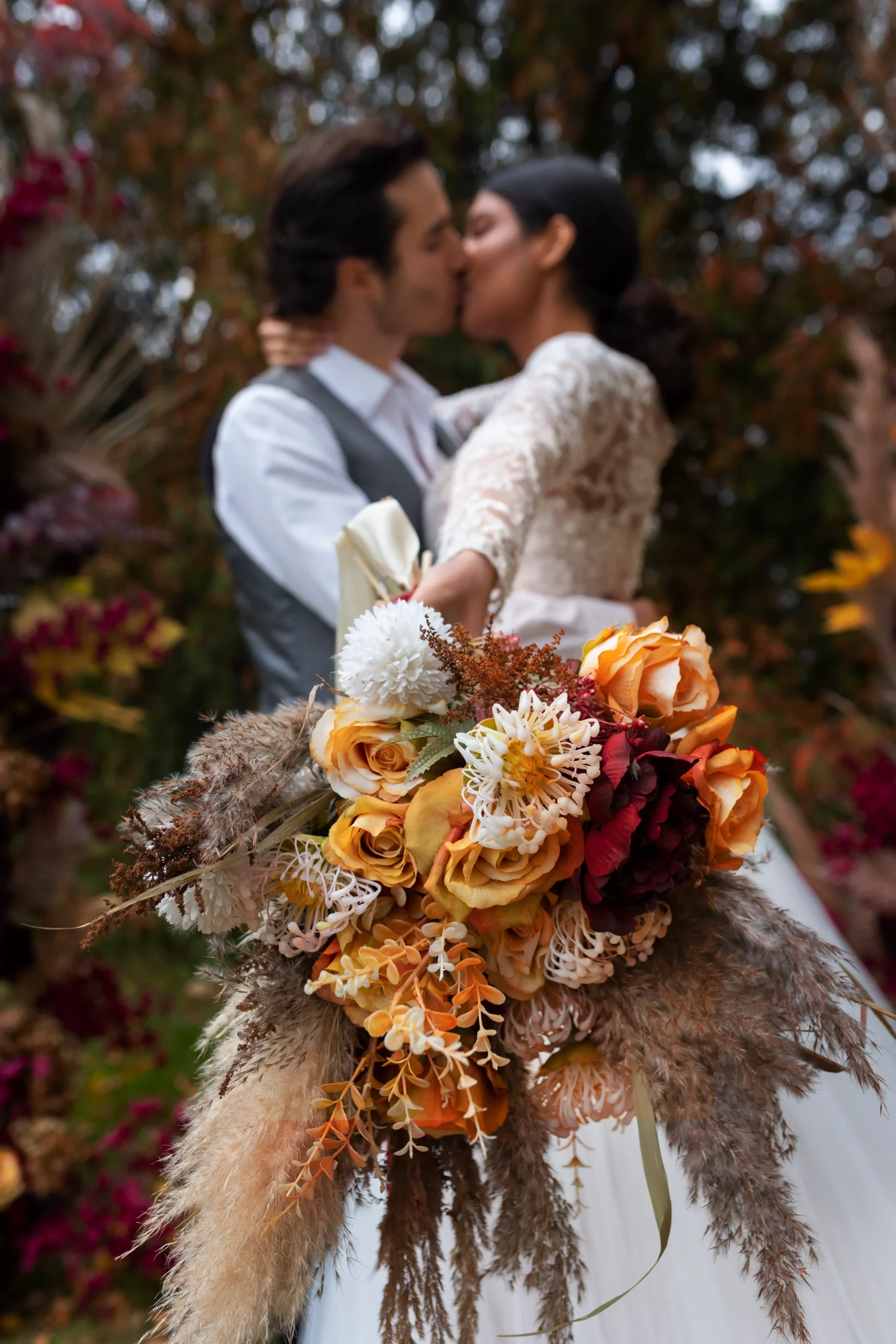 Pareja de novios en su boda, besándose mientras la chica muestra su ramo de flores delante de la cámara con el brazo estirado